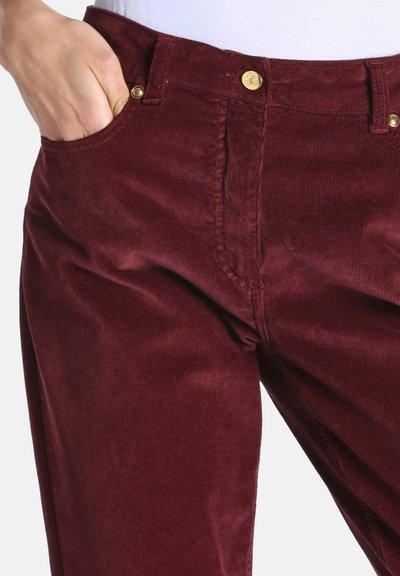 Personne portant un pantalon en velours côtelé rouge foncé avec la main dans la poche avant et des détails de boutons dorés sur la taille et la poche.
