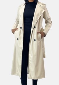 Beige trenchcoat met een gladde satijnen textuur, dubbele rij knopen, grote zwarte knopen en steekzakken. Op maat gemaakt ontwerp, knielengte.