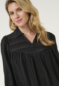 Melna krekls ar apkakli, ar vertikālām svītrām, viegls audums un brīvs griezums ar aizsēdējošām detaļām pie krūšu daļas.