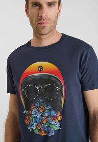 Hombre vistiendo una camisa azul marino con un gráfico de un casco negro y gafas, rodeado de rosas azules y mariposas naranjas.