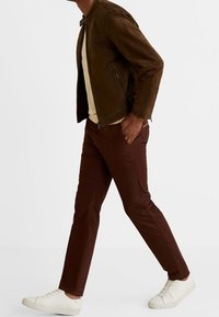 Brązowa zamszowa kurtka o dopasowanym kroju, beżowa koszula pod spodem, ciemne brązowe spodnie slim-fit i białe sneakersy; swobodny strój w ruchu.