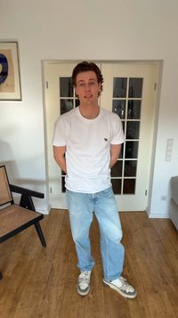 Fiatal férfi fehér pólóban, világoskék farmerben és sportcipőben, egy szobában áll, mögötte fa padló és üveges, dupla ajtók.
