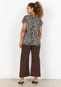 Schwarzes und braunes, zebra-musteriertes Kurzarmshirt, kombiniert mit braunen, weit geschnittenen Hosen und schwarzen, offenen Absatzschuhen, vor einem weißen Hintergrund.