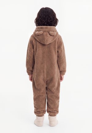 Enfant aux cheveux bouclés foncés, debout de dos, portant une grenouillère marron duveteuse avec capuche, petites oreilles sur la capuche, et des chaussons blancs.