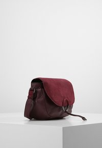 Bordeauxrode leren crossbodytas met een suède klep, ronde metalen details, zachte structuur en een trekkoordsluiting; compact en gestructureerd model.
