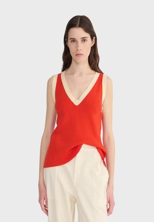 Frau mit langen braunen Haaren, die ein ärmelloses rotes Strickoberteil mit beiger Borte und weite weiße Hosen trägt, vor einem einfarbigen hellen Hintergrund.