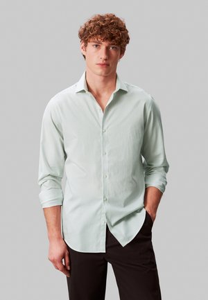 Ung mann med krøllete hår iført en lysegrønn stripete skjorte med knapper og svarte bukser, stående med den ene hånden i lommen.