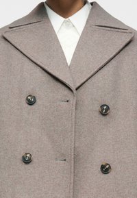 Κοντινό πλάνο ενός γκρι διπλόφτερου παλτό από μαλλί με φαρδιές πέτες και τέσσερα σκουρόχρωμα μαρμάρινα κουμπιά, φορεμένο πάνω από ένα λευκό πουλόβερ με γιακά.