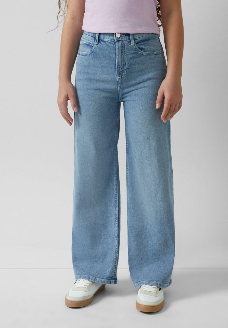 Personne portant un jean large bleu clair, des baskets blanches avec des semelles en gomme et un haut violet clair, debout devant un fond uni.