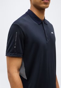 Άνδρας που φορά μπλε ναυτικό πόλο με το λογότυπο JL στο στήθος και το κείμενο "J.LINDEBERG" στο αριστερό μανίκι.