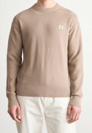 Béžový pletený svetr s texturou, s žebrovanými manžetami a lemem, nošený s bílými kalhotami, s malou obdélníkovou logovou nášivkou na hrudi.