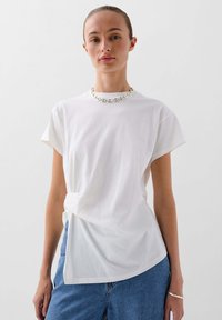 T-shirt bianca in cotone con maniche corte, caratterizzata da un dettaglio frontale attorcigliato. Abbinata a jeans azzurri chiari. Design semplice e casual.