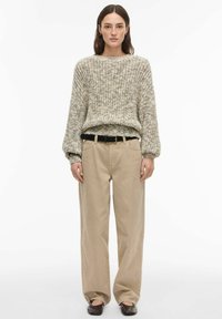 Pull en tricot épais beige avec des touches multicolores subtiles, assorti à un pantalon beige à jambes larges et une ceinture noire. Chaussures plates lisses.