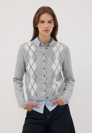 Mujer joven con cárdigan gris de rombos sobre camisa azul con rayas y botones, con las manos en los bolsillos de pantalones oscuros frente a un fondo liso.