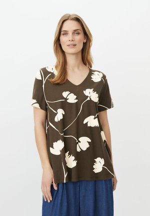 Kvinde iført en brun kortærmet V-hals top med store hvide blomsterprint, kombineret med løstsiddende blå bukser mod en ensfarvet baggrund.