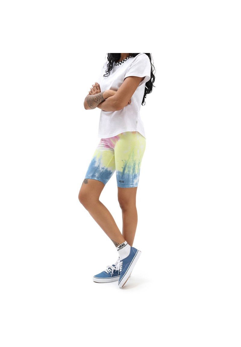 Personne portant un t-shirt blanc Vans, un cuissard de cyclisme tie-dye jaune-bleu, des chaussettes blanches Vans et des baskets bleues Vans, debout les bras croisés.