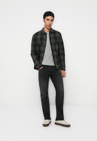 Chemise en flanelle à carreaux verts et noirs superposée à un pull gris, accompagnée de jeans foncés et de baskets blanches. Tissu lisse, coupe décontractée.