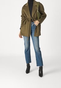 Oliivinvihreä villasekoitetakki, jossa on vyötärönauhavyö ja kaksi etutaskua, yhdistetty vaaleansinisiin liehuviin farkkuihin ja mustiin, teräväkärkisiin nilkkureihin.