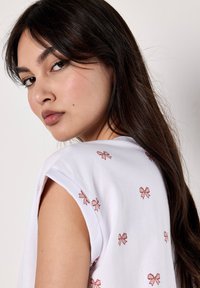 Una mujer con cabello largo y oscuro mira por encima de su hombro, vestida con una blusa blanca adornada con pequeños lazos rosas. Una suave luz natural resalta sus rasgos.
