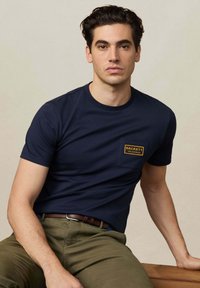 Camiseta de algodón azul marino con un pequeño bolsillo en el pecho que lleva una etiqueta amarilla "Hackett Velospeed". Diseño sencillo de cuello redondo.