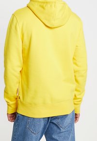 Sudadera con capucha amarilla de mezcla de algodón, con puños y dobladillo acanalados. Textura suave; etiqueta con logotipo cerca de la parte inferior en el lado izquierdo.