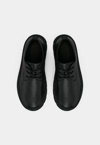 Sapatos de couro preto com biqueiras arredondadas, textura lisa e solas planas. Apresentam seis ilhós para atacadores e uma marca subtil na língua.