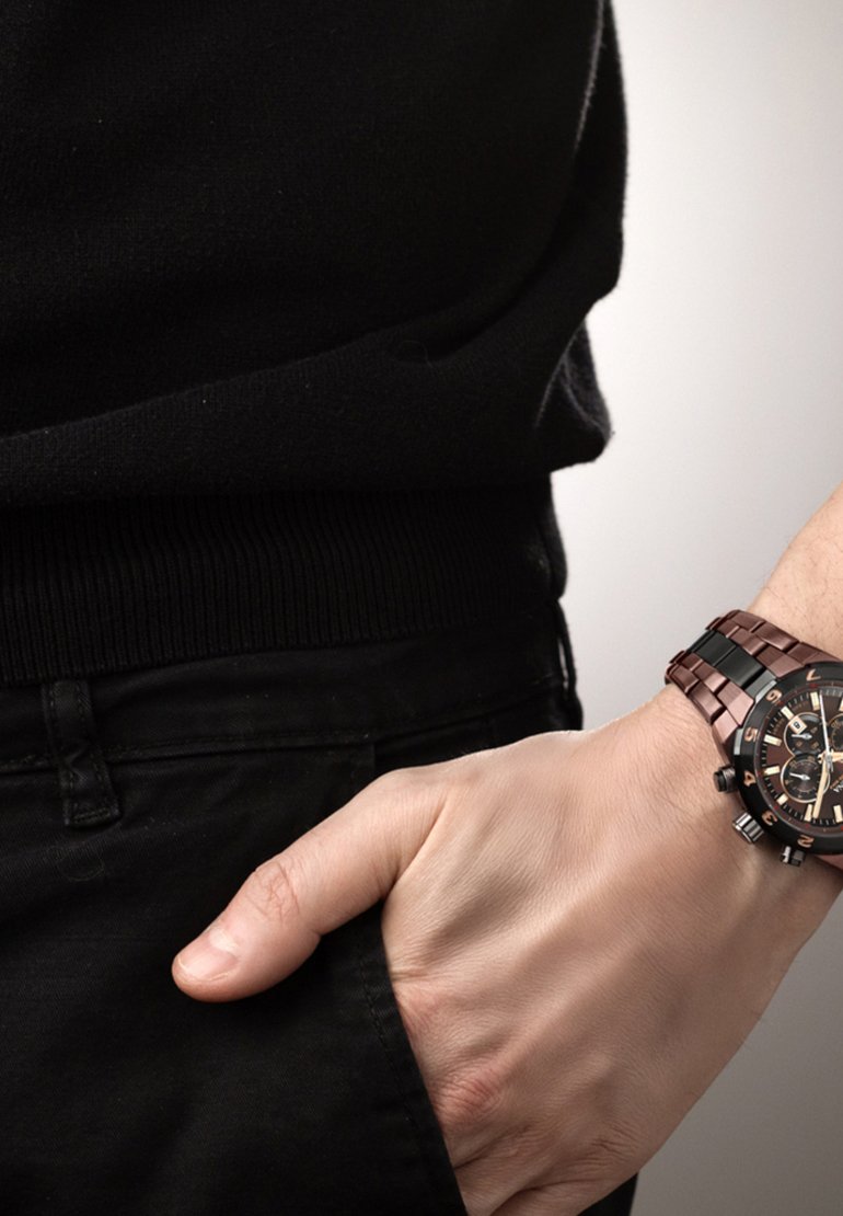 Bruine metalen horloge met een zwarte wijzerplaat, voorzien van een gepolijste armband en lichtgevende wijzers. Draagt het om een pols, samen met zwarte kleding.