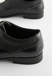 Zapatos de cuero negro con detalles de tela texturizada, diseño curvado y tacón bajo. Presenta sutiles costuras a lo largo de los bordes.