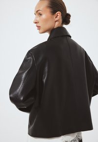 Nainen, jolla on hiukset nutturalla, pukeutuneena löysään mustaan nahkatakkiin ja valkoiseen pitsireunukseen vaatetukseen, katsomassa vasemmalle yksinkertaista taustaa vasten.