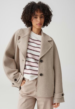 Donna con capelli ricci che indossa un cappotto di lana beige sopra un maglione bianco e rosa a righe e pantaloni beige chiaro, in piedi con una mano in tasca.