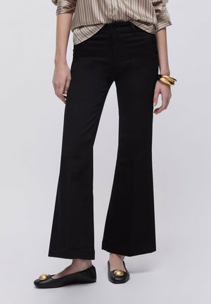 Frau trägt schwarze Schlaghose, schwarze Ballerinas mit goldenen Akzenten, gestreiftes Hemd und goldene Armbänder am Handgelenk vor schlichtem Hintergrund.