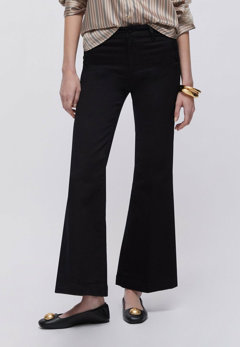Femme portant un pantalon évasé noir, des ballerines noires avec des accents dorés, une chemise rayée et des bracelets dorés au poignet, sur un fond uni.
