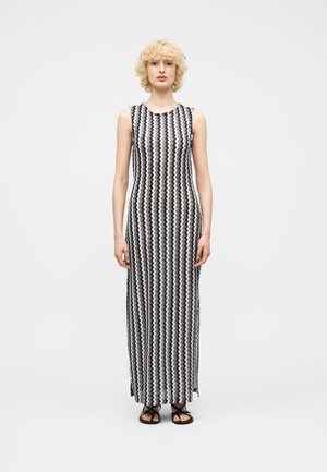 Mujer con cabello rubio rizado, vestida con un vestido largo sin mangas con patrón de zigzag en blanco y negro, y sandalias negras, de pie sobre un fondo blanco.