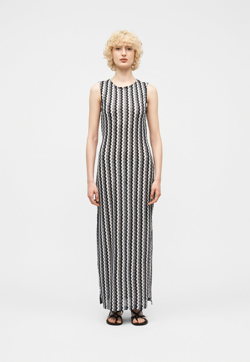 Femme aux cheveux blonds bouclés portant une robe maxi sans manches à motif zigzag noir et blanc et des sandales noires, debout droite sur fond blanc.