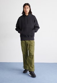 Schwarzer Kapuzenpullover mit Fronttasche und Kordelzug; kombiniert mit olivgrünen Cargo-Hosen im geraden Schnitt und verstellbarem Bund.