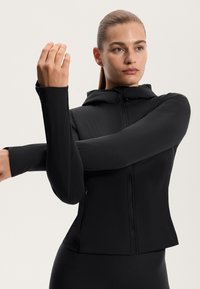 Černá sportovní bunda s předním zipem a kapucí. Má dlouhé rukávy s otvory na palce, úzký střih a hladký, pružný materiál.