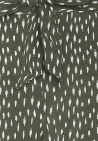 Olijfgroen stof met een witte onregelmatige penseelstreekpatroon, met een gestrikte strikdetail bij de halslijn. Zacht, textuurrijk materiaal.