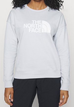 Person iført en lysegrå langærmet sweatshirt med hvidt The North Face-logo og sorte bukser, stående foran en ensfarvet baggrund.