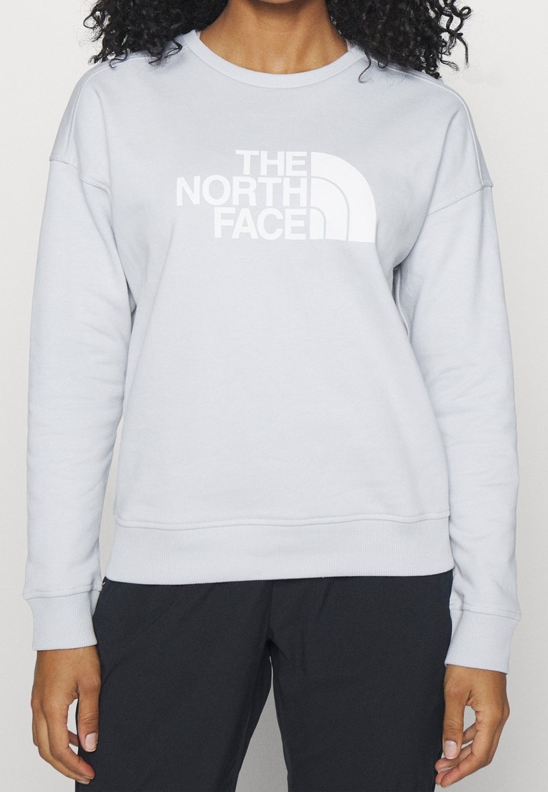 Personne portant un sweat-shirt gris clair à manches longues avec le logo blanc The North Face et un pantalon noir, debout devant un fond uni.