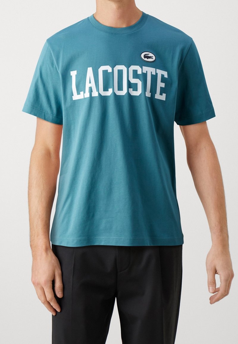 Tyrkysové bavlněné tričko s kulatým výstřihem, na levé straně s bílým textem "LACOSTE" a nášivkou loga. Krátké rukávy, volný střih.