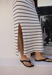 Gestreiftes, weißes Maxikleid mit marineblauen Linien und seitlichem Schlitz; kombiniert mit schwarzen Flip-Flops auf einer weißen Fläche vor einer verwitterten Tür.