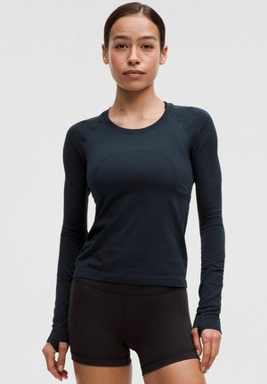 Kvinde iført en tætsiddende sort langærmet top og sorte atletikshorts, stående foran en ensfarvet lys baggrund.