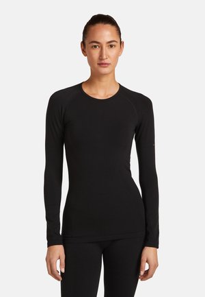 Nainen seisoo kasvot eteenpäin, pukeutuneena istuvaan mustaan pitkähihaiseen yläosaan ja vastaaviin mustiin alaosiin yksiväristä vaaleaa taustaa vasten.