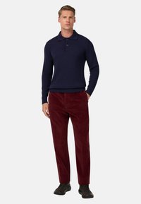 Navy-blaues Strick-Polohemd mit Kragen und zwei Knöpfen, kombiniert mit burgunderfarbenen Cordhosen und dunklem Schuhwerk, was ein kontrastierendes Outfit ergibt.