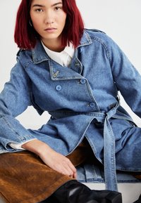 Trench coat in denim con bottoni doppi, vita con cintura e tessuto a contrasto sotto in suede marrone. Modella seduta su una superficie neutra.
