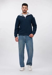 Man staat in een marineblauwe half-zip sweater, lichtblauwe spijkerbroek en witte sneakers tegen een plain witte achtergrond.