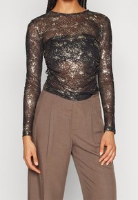 Durchsichtige schwarze Bluse mit langen Ärmeln, ausgestattet mit goldenen metallischen Sprenkeln und einem gerafften Design an der Vorderseite, kombiniert mit hellbraunen hoch taillierten Hosen.