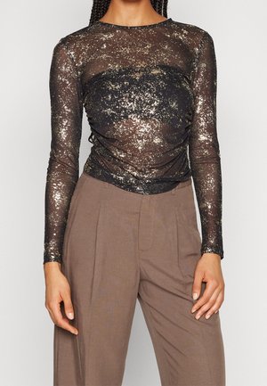 Durchsichtige schwarze Bluse mit langen Ärmeln, ausgestattet mit goldenen metallischen Sprenkeln und einem gerafften Design an der Vorderseite, kombiniert mit hellbraunen hoch taillierten Hosen.