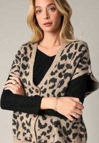Gilet beige avec un imprimé léopard noir, fermeture à boutons, motif tricoté texturé. Porté par-dessus un pull noir, avec des bagues et un bracelet visibles.