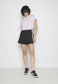 Pantaloni sportivi neri con strisce bianche sui lati, abbinati a una t-shirt rosa chiaro con un logo tonale. Il modello indossa occhiali da sole neri e scarpe da ginnastica.